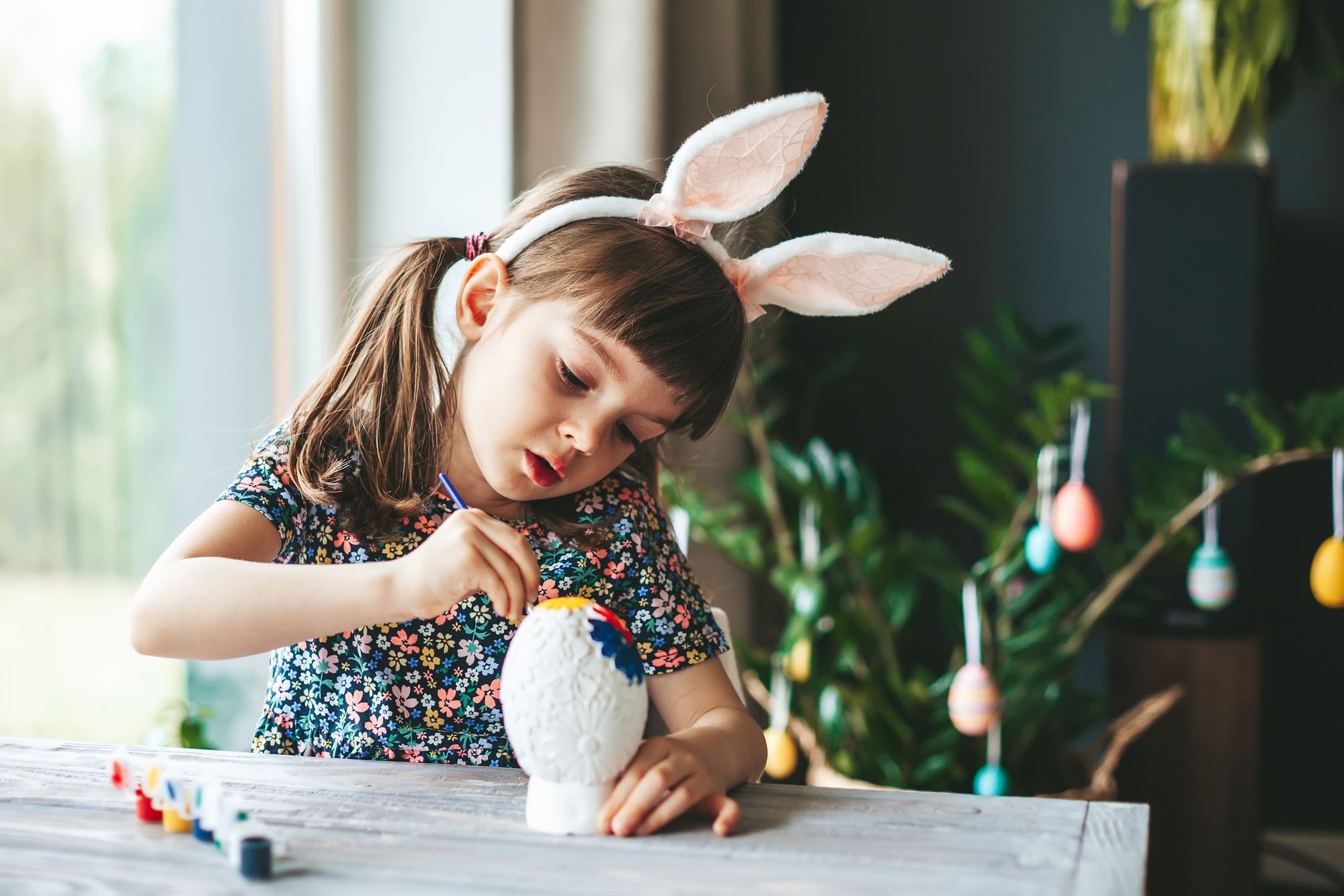 Cute little girl with bunny ears painting Easter egg with paintbrush